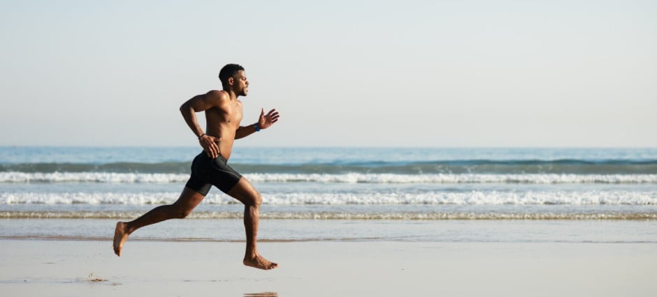 Foto de homem correndo descalço na areia, em frente ao mar