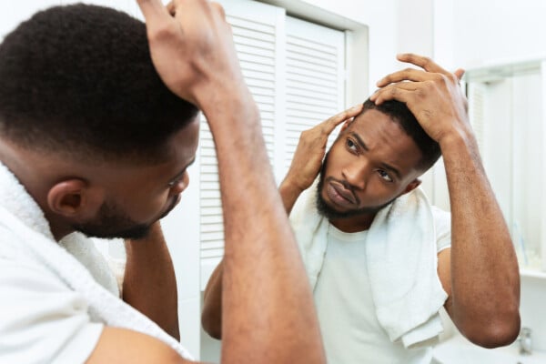 Homem negro, veste camiseta branca, se olha no espelho checando queda de cabelo