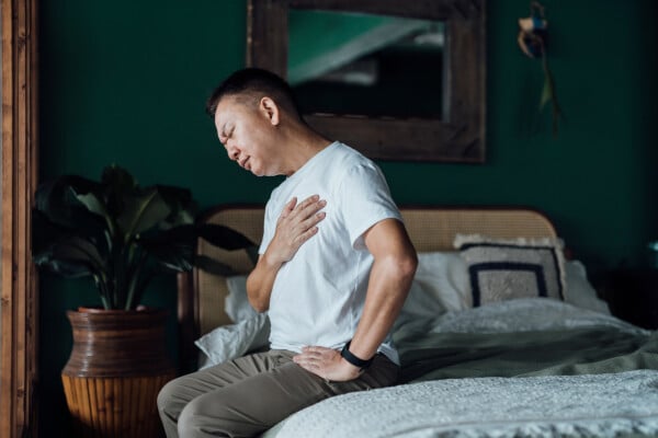 Homem asiático sentado na cama com a mão no peito