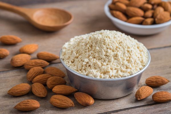 Farinha de amêndoas servida em uma tigela, ao lado de várias amêndoas sobre uma mesa de madeira