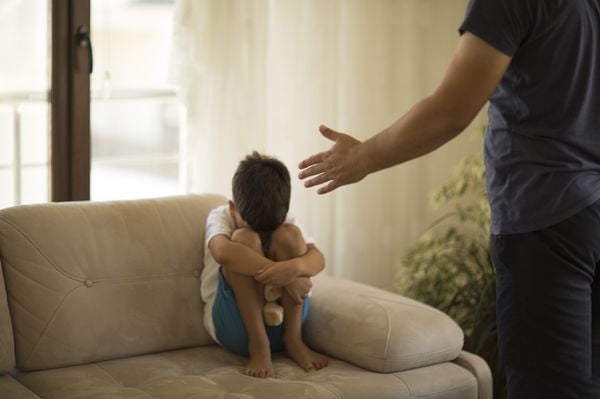 Menino retraído no sofa, com a cabeça baixa entre os joelhos, enquanto escuta um adulto