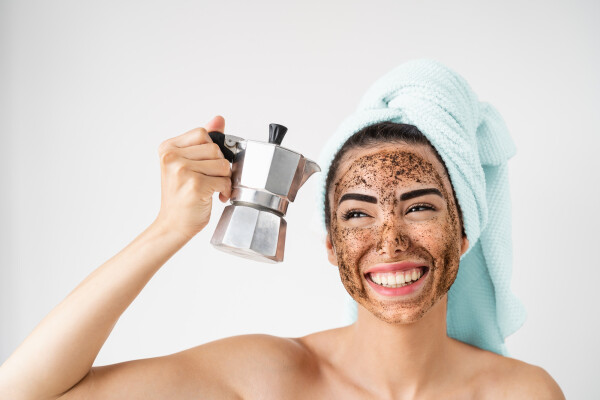 Mulher branca com uma toalha no cabelo e borra de café no rosto segurando um bule de café