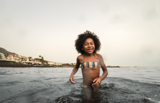 Criança afro-americana, sorridente, de biquíni de listras coloridas, brincando no mar, com a água até a região da cintura. Ao fundo, casas e palmeiras altas.
