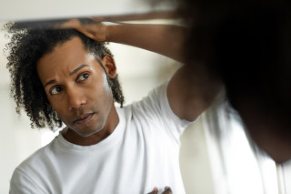 Homem negro, vestido de camiseta branca, com cabelos cacheados compridos olhando no espelho, preocupado com entradas típicas de calvície