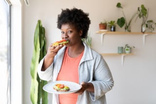 Mulher comendo um pão com folhas verdes e tomates enquanto segura um prato