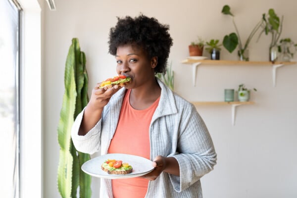 Mulher comendo um pão com folhas verdes e tomates enquanto segura um prato