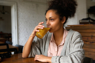 mulher sentada com um braço apoiado em mesa de madeira enquanto bebe um copo de suco de laranja