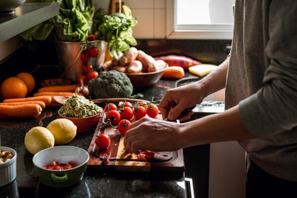 Recorte de imagem de mulher cortando tomates em uma tábua de madeira em cima de uma pia; há vários legumes e frutas ao redor