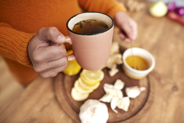 Mulher segurando caneca com chá e, ao fundo, cabeças de alho, limão em rodelas e pote com mel
