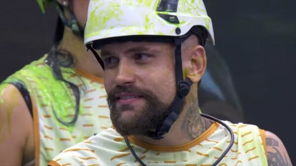 Vinícius Rodrigues durante participação na prova do líder do BBB24; ele veste colete branco com listras laranjas e capacete branco com manchas verdes
