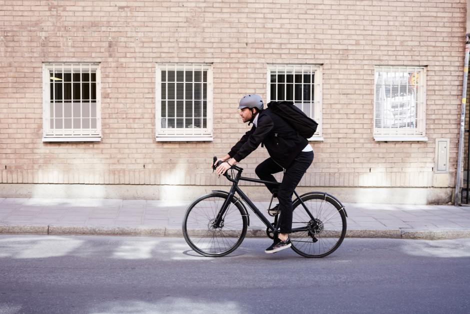 Homem andando de bicicleta em rua usando uma mochila, capacete e roupa preta