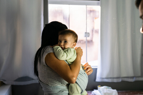 Mãe, de cabelos lisos e pretos, veste camiseta cinza e está abraçada com seu bebê