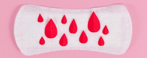 Foto de um absorvente, em um fundo rosa, com manchas ilustrativas de sangue, feitas de papel