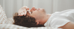 Homem jovem deitado em uma cama com a mão na testa, em sinal de confusão
