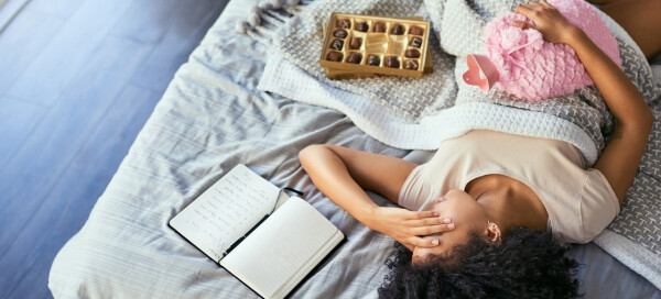 Mulher deitada na cama, ao lado de um caderno e de uma caixa de bombons, segura uma bolsa térmica na região do abdômen. Ela está com sintomas de TPM.