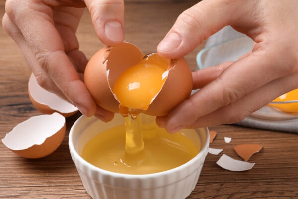 ovo sendo quebrado dentro de pote branco, que está em cima de mesa de madeira