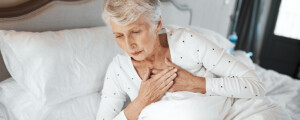 Mulher idosa deitada na cama com a mão no coração, sinalizando desconforto