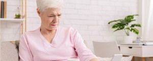 Mulher idosa, pele branca e cabelos brancos curtos, veste blusa rosa e segura um celular ao longe, com dificuldade de enxergar devido à hipermetropia