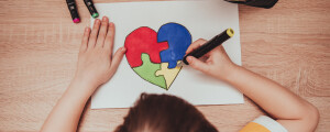 Imagem vista de cima de um garoto loiro e branco, vestindo camiseta amarela e branca listrada, desenhando um quebra-cabeça colorido em formato de coração, em referência ao símbolo do autismo