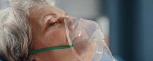 Imagem de uma senhora em ambiente hospitalar, deitada em uma cama usando máscara de oxigênio