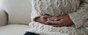 Mulher sentada no sofá com as mãos apoiadas na região do abdômen; dor abdominal é um dos sintomas de endometriose intestinal