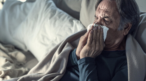 Homem deitado na cama com cobertor em volta dos ombros enquanto assoa o nariz