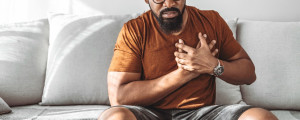 Homem negro aflito com camiseta marrom, sentado em um sofá cinza e com as mãos no peito, na região do coração.