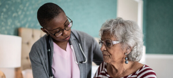 Médica, de pele negra e cabelos raspados escuros, atende uma senhora idosa branca de cabelos curtos e grisalhos com Alzheimer