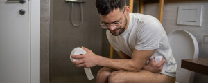 Homem sentado no vaso sanitário enquanto segura um rolo de papel higiênico