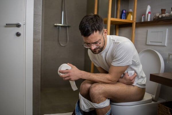 Homem sentado no vaso sanitário enquanto segura um rolo de papel higiênico