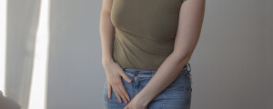 Recorte de imagem de mulher vestindo calça jeans e camiseta verde escuro, com as mãos sobre a região da virilha, sentindo dor