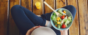 Mulher grávida sentada no chão com um prato de salada com folhas e vegetais em uma das mãos