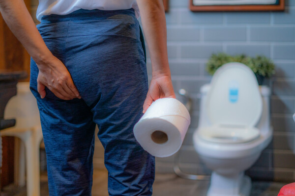 Homem com a mão nos glúteos segurando um papel higiênico de frente para um vaso sanitário