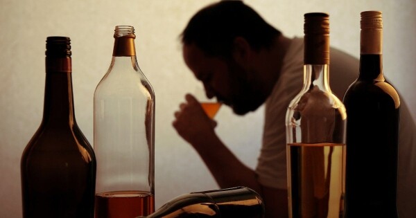 Homem bebendo um copo com bebida alcóolica. Quatro garrafas de bebida alcóolica em uma superficie na frente do homem.