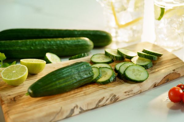 Imagem de um pepino, com uma metade fatiada e outra completa, em uma tabua de madeira, onde alguns limãos também estão dispostos.