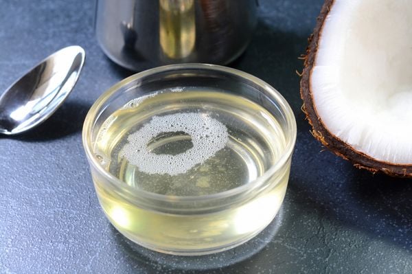 Recipiente transparente com óleo de coco em cima de mesa escura. Ao lado, a metade de um coco.