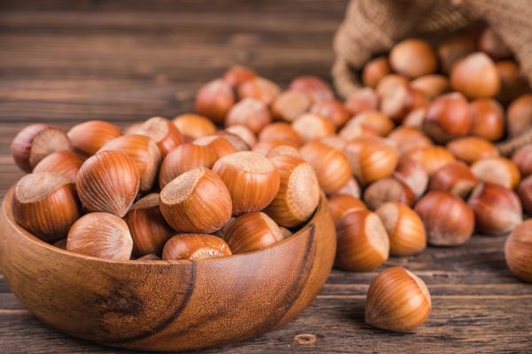 Na Imagem algumas avelãs espalhadas sobre uma mesa de madeira e outras em um pote, também de madeira