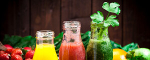 Foto com três garrafas de vidro com sucos detox; ao fundo, frutas e verduras espalhadas, como morango, laranja, limão e couve