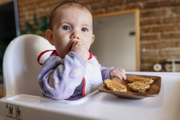 Bebê sentando em uma cadeirinha enquanto come uma paçoca de amendoim. Na frente dele, em cima da cadeirinha, há um recipiente de madeira com mais três paçocas.