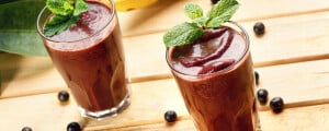 Dois copos de suco de açaí decorados com folhas de hortelã em cima de mesa de madeira com o açaí em fruta espalhado