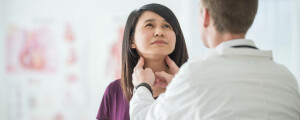 Mulher sendo analisada por médico, que toca com os dedos o pescoço da mulher.