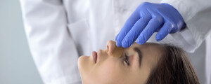 Médico com luvas azuis coloca um curativo em nariz de paciente do sexo feminino, com cabelos escuros