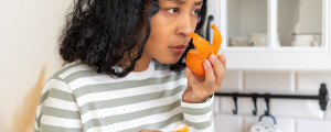Mulher de pele em tonalidade média com cabelos ondulados pretos usando blusa listrada em cinza e branco cheira cascas de laranja em uma cozinha