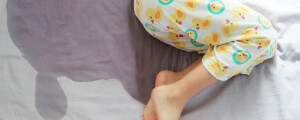 Foto aproximada de pernas de criança deitada com mancha de xixi na cama ao lado