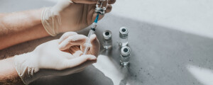 Imagem de uma mão usando luvas de látex segurando uma siringa e um pote de medicamento