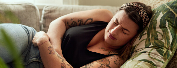 Mulher branca, com tatuagens no braço e no ombro, deitada em sofá veste regata preta e calça jeans, enquanto abraça a região abdominal, com dores típicas de endometriose