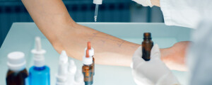 Imagem ampliada do antebraço de uma pessoa em cima de uma mesa, enquanto um médico pinga um medicamento alérgeno para um teste de alergia