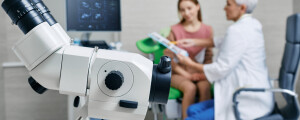 aparelho de colposcopia (colposcópio) em foco. atrás dele, uma paciente e sua dermatologistas estão sentadas olhando os resultados do exame
