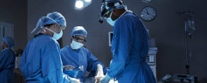 três cirurgiões fazendo uma cirurgia em um paciente deitado em maca em uma sala de cirurgia