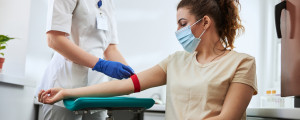 Mulher branca veste camiseta clara e está sentada esperando para fazer exame de sangue, enquanto enfermeira, vestida de branco e com pele branca, amarra um elástico no braço da paciente